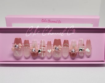 May include: A set of 10 pink and white press-on nails with different designs, including bows, hearts, and plaid patterns. The nails are displayed in a pink box with the text "Cake Channel Co."