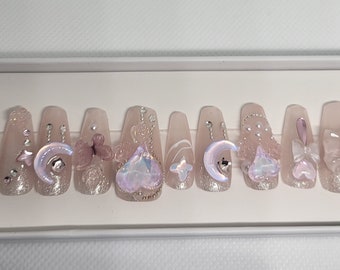 May include: A set of ten long, coffin-shaped, light pink press-on nails with glitter and various embellishments, including moons, stars, hearts, butterflies, and bows.