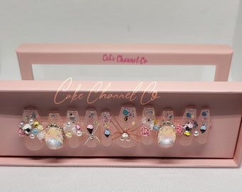May include: A set of ten clear press-on nails with various colorful designs and embellishments, including flowers, glitter, and a small ice cream cone. The nails are displayed in a pink box with the text "Cake Channel Co."