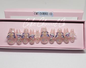 May include: A set of 10 pink press-on nails with butterfly designs and rhinestones. The nails are displayed in a pink box with the text "Cake Channel Co."