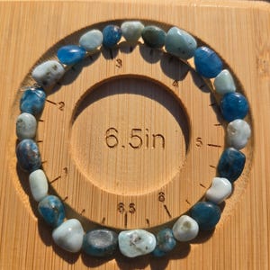 May include: A blue and white gemstone bracelet, approximately 6.5 inches in diameter, composed of various shades of blue and white stones. The bracelet is displayed on a wooden measuring board with the text "6.5in".
