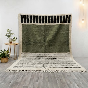 May include: A rectangular area rug featuring a large central olive green block framed by cream-coloured borders. The top border displays thick black vertical stripes, while the bottom border contains numerous thin horizontal black lines and fringe.
