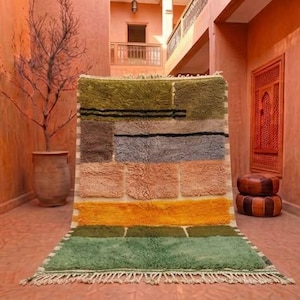 May include: A rectangular wool rug featuring a geometric block pattern in shades of olive green, charcoal grey, muted orange, and bright yellow. The rug has a thick, high-pile texture with horizontal black lines and a fringed edge at the bottom.