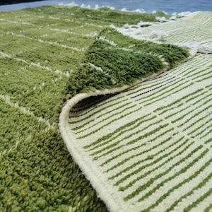 May include: A close-up of a green and white rug with a textured, shaggy pile. The rug features a pattern of green lines and a rolled-up edge, revealing the underside with a striped design. The rug is a soft, decorative home textile.