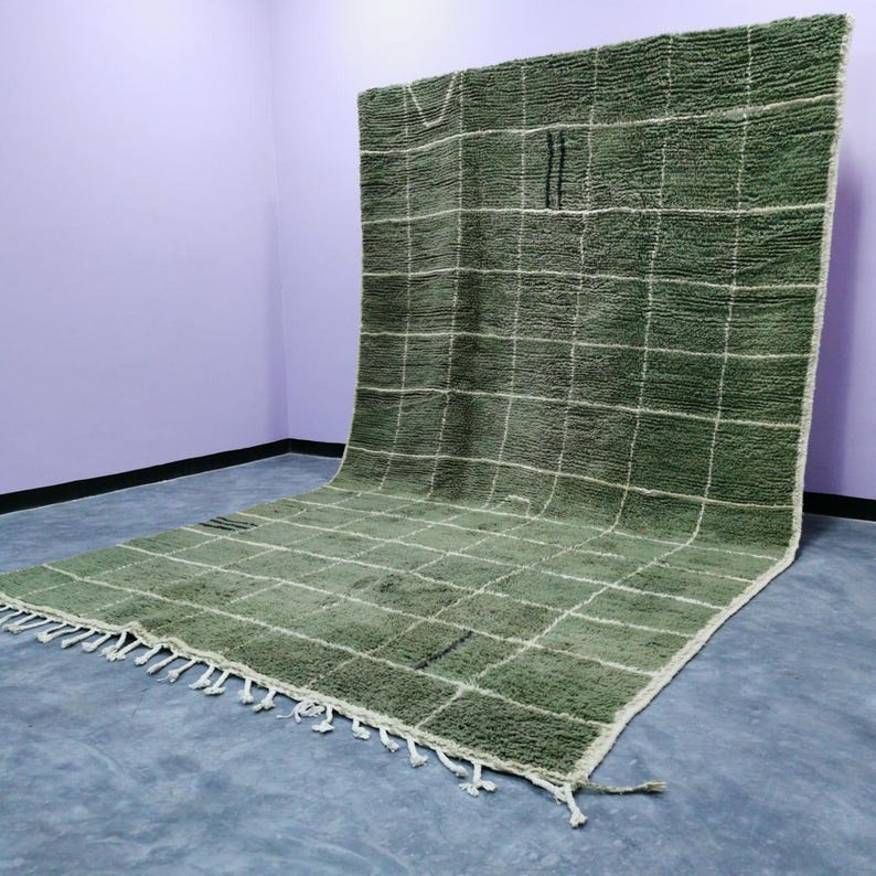 May include: A large, olive green rug with a grid pattern of white lines. The rug has a thick, textured pile and a fringe along one edge. The rug is displayed against a purple wall and a gray floor.