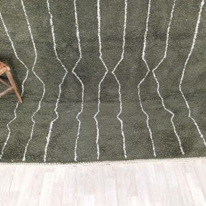 May include: Olive green shag rug with white geometric lines. The rug has a thick pile and a textured appearance. A wooden chair with a woven seat and a leather pouf are in the background.