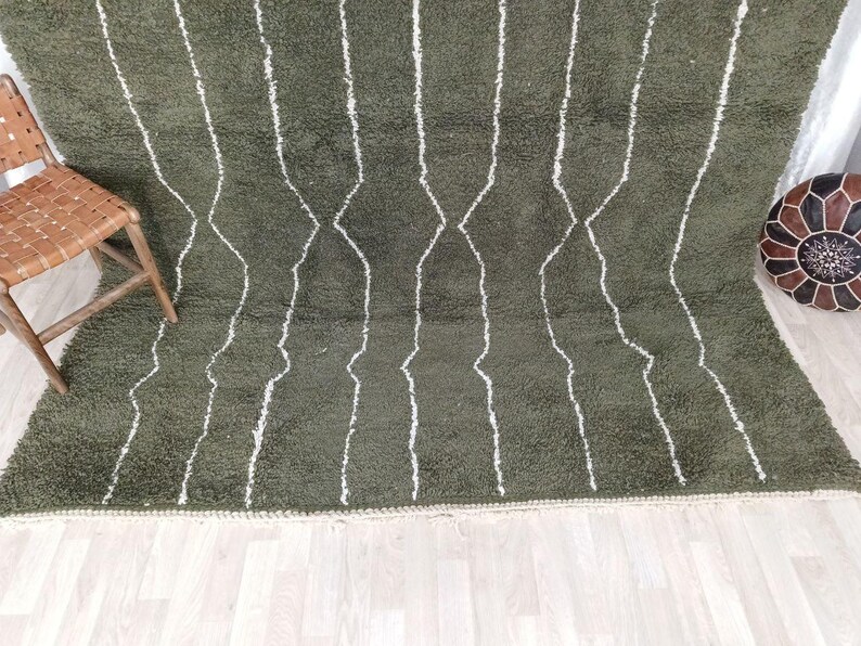 May include: Olive green shag rug with white geometric lines. The rug has a thick pile and a textured appearance. A wooden chair with a woven seat and a leather pouf are in the background.