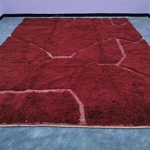 May include: A large, rectangular, burgundy rug with a geometric design. The rug features a plush, textured surface with lighter-colored, linear accents that define the pattern. The rug is placed on a gray surface.