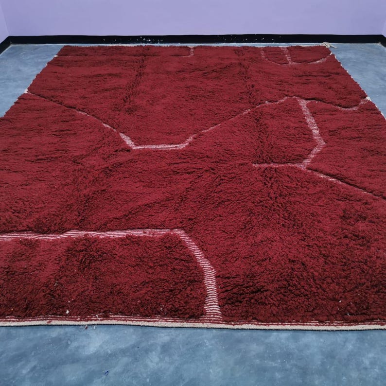 May include: A large, rectangular, burgundy rug with a geometric design. The rug features a plush, textured surface with lighter-colored, linear accents that define the pattern. The rug is placed on a gray surface.