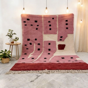 K&ouml;nnte beinhalten: Ein gro&szlig;er, rechteckiger Teppich mit geometrischem Design. Der Teppich hat ein rosa-cremefarbenes Farbschema mit schwarzen quadratischen Akzenten. Der untere Rand ist dunkler rot und der Teppich hat einen Fransenrand. Der Teppich wird vor einer wei&szlig;en Wand ausgestellt.