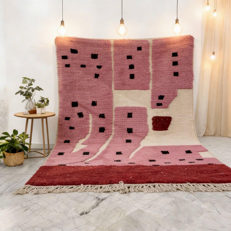 K&ouml;nnte beinhalten: Ein gro&szlig;er, rechteckiger Teppich mit geometrischem Design. Der Teppich hat ein rosa-cremefarbenes Farbschema mit schwarzen quadratischen Akzenten. Der untere Rand ist dunkler rot und der Teppich hat einen Fransenrand. Der Teppich wird vor einer wei&szlig;en Wand ausgestellt.