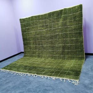 May include: A large, olive green rug with a grid pattern of white lines. The rug has a textured surface and a fringe along one edge. The rug is displayed against a purple wall and a gray floor.
