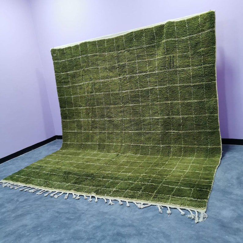 May include: A large, olive green rug with a grid pattern of white lines. The rug has a textured surface and a fringe along one edge. The rug is displayed against a purple wall and a gray floor.