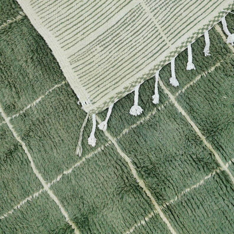 May include: A close-up of a green and cream-colored rug with a geometric pattern. The rug has a plush texture with a grid design in cream-colored lines. The top section of the rug has a striped pattern and white tassels.
