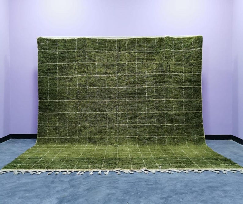 May include: A large, olive green rug with a grid pattern of white lines. The rug has a shaggy texture and a fringe along the bottom edge. The rug is displayed against a purple wall and blue floor.