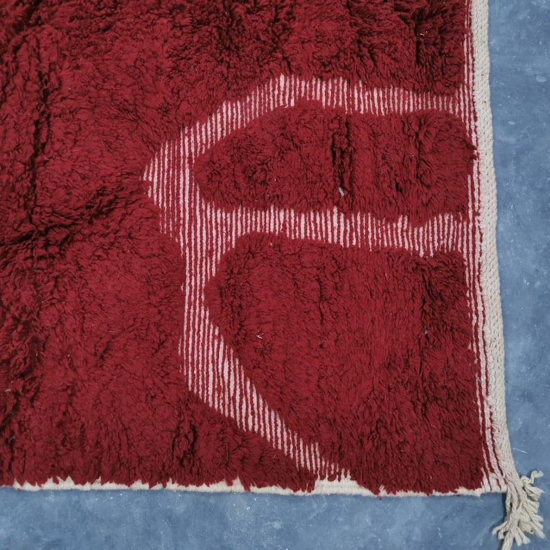 May include: A close-up of a burgundy rug with a white geometric design. The rug has a shaggy texture and a white fringe along the edge. The design is composed of white vertical lines and shapes.