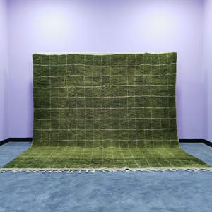 May include: A large, olive green rug with a grid pattern of white lines. The rug has a thick, textured pile and a fringe along the bottom edge. The rug is displayed against a purple wall and a gray floor.