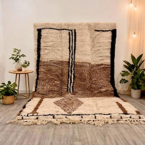 May include: A large rectangular area rug featuring a thick, shaggy pile in cream, tan, and brown tones. The design includes bold black vertical lines, a central diamond-shaped motif, and a fringed edge along the bottom of the textile.