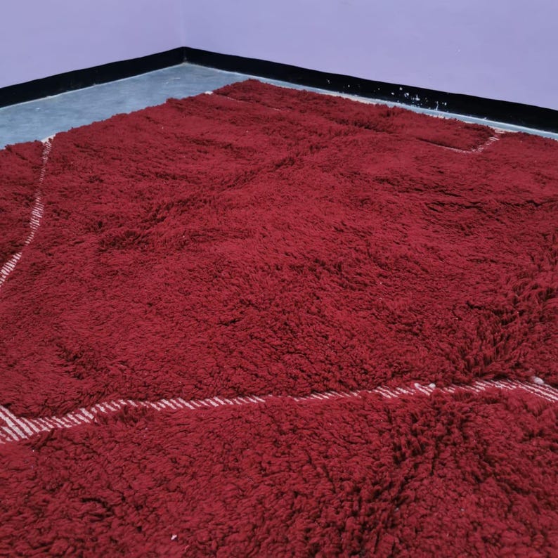 May include: A plush, rectangular rug in a deep burgundy color. The rug has a thick, textured pile and a white and red striped border. The rug is placed in a corner of a room with purple walls.