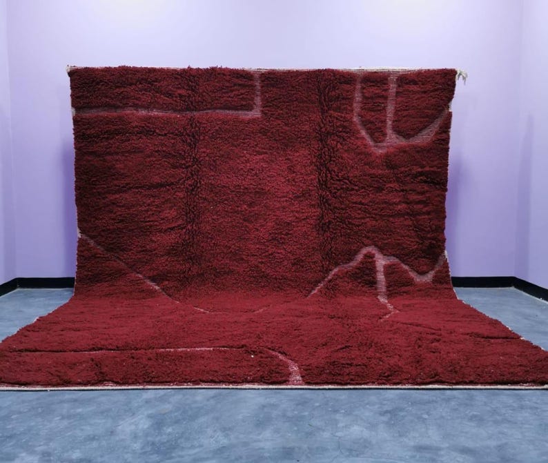 May include: A large, burgundy-colored rug with a shaggy texture. The rug features a geometric design in a lighter shade, creating a contrast. The rug is displayed against a purple wall and a gray floor.