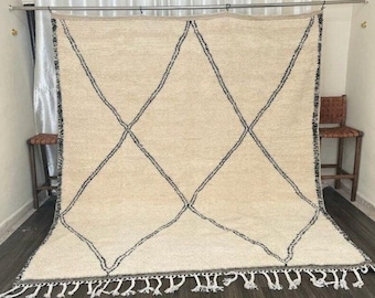May include: Cream-colored rug with a large black diamond pattern. The rug has a fringe along the bottom edge. The rug is displayed in a room with a dark wood floor and a neutral-colored wall.