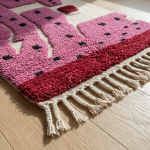 K&ouml;nnte beinhalten: Ein rechteckiger Teppich mit geometrischem Design. Der Teppich hat einen hellbeigen Hintergrund mit rosa und roten Formen, akzentuiert mit schwarzen Punkten. Der untere Rand hat cremefarbene Fransen.