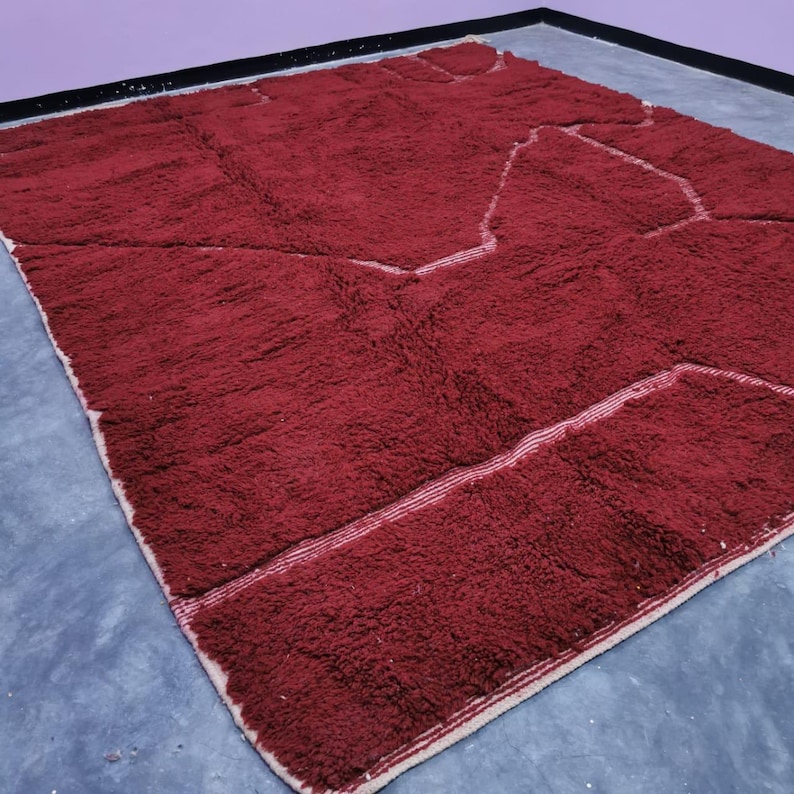 May include: A large, rectangular, burgundy-colored rug with a geometric design. The rug has a thick, textured pile and a cream-colored border with a thin white stripe. The design features lighter burgundy shapes outlined with white lines.