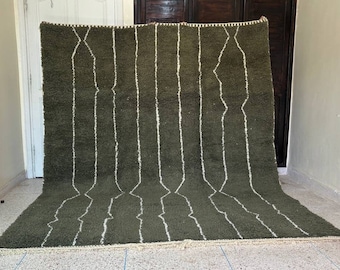 May include: Olive green rug with vertical white geometric patterns. The rug has a shaggy texture and a cream-colored fringe along the bottom edge. The design features a series of white lines that create a repeating pattern across the surface.