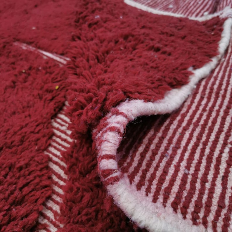 May include: A close-up of a red and white rug. The rug has a thick, textured pile in a deep red color. The edges are finished with a white border. The underside of the rug reveals a striped pattern in red and white.
