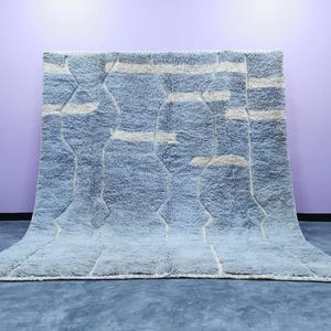 Könnte beinhalten: Ein großer, rechteckiger Teppich mit einem hellblauen und cremefarbenen geometrischen Muster. Der Teppich hat ein weiches, strukturiertes Aussehen und wird vor einer lila Wand präsentiert. Das Design weist vertikale und horizontale Linien auf.