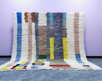 May include: A rectangular, off-white rug with a geometric pattern. Vertical stripes in blue, brown, yellow, and pink are interspersed across the rug. The rug has a textured, woven appearance and a fringed edge.
