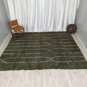 May include: An olive green rug with white, wavy lines. A wooden chair with a woven seat and back is in the upper left corner. A decorative, round, patterned cushion sits in the upper right corner. The rug is on a light wood floor.