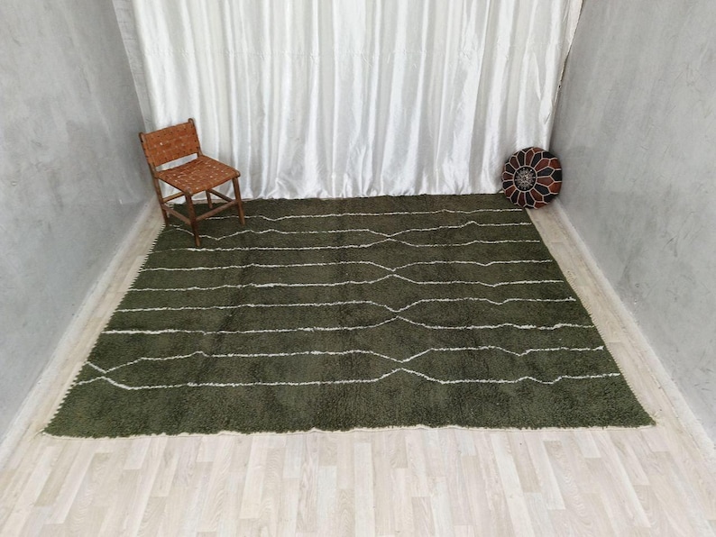 May include: An olive green rug with white, wavy lines. A wooden chair with a woven seat and back is in the upper left corner. A decorative, round, patterned cushion sits in the upper right corner. The rug is on a light wood floor.