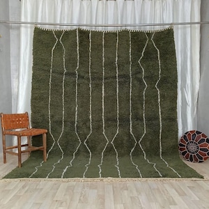 May include: An olive green Moroccan rug featuring a white geometric diamond and line design. The rug is displayed against a white curtain backdrop, with a wooden chair and a patterned ottoman in the room. The rug is a floor covering.