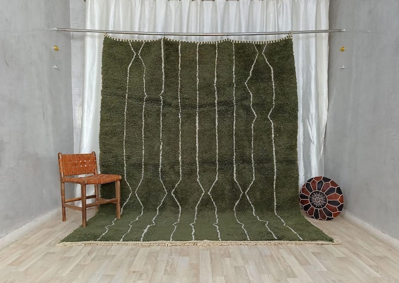 May include: An olive green Moroccan rug featuring a white geometric diamond and line design. The rug is displayed against a white curtain backdrop, with a wooden chair and a patterned ottoman in the room. The rug is a floor covering.