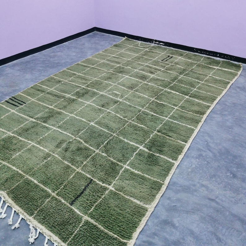 May include: A rectangular, olive green rug with a grid pattern of white lines. The rug has a shaggy texture and a fringe along the bottom edge. The rug is placed on a gray surface.