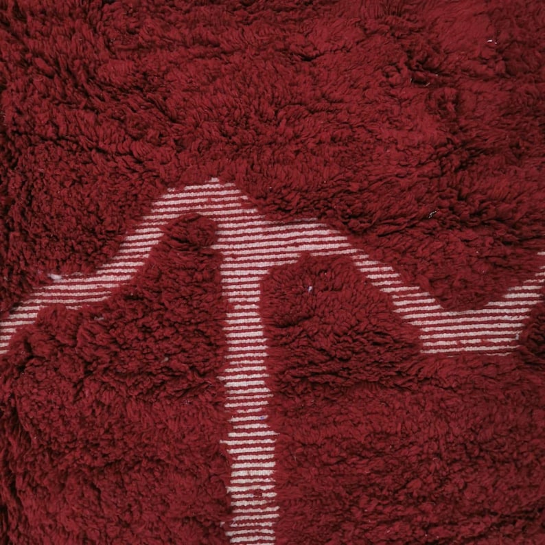 May include: A close-up of a plush, burgundy rug with a white, geometric design. The rug has a thick, textured pile, and the white design is made up of thin, parallel lines. The overall aesthetic is warm and inviting.