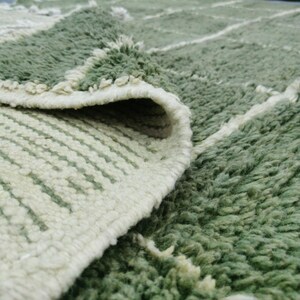May include: Close-up of a green and white rug with a geometric pattern. The rug has a thick, textured pile and a folded edge, revealing the underside with a striped design. The color palette is primarily green and white.