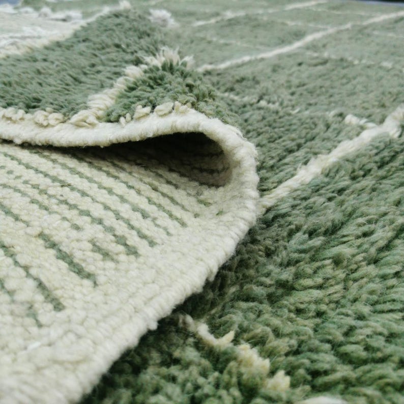 May include: Close-up of a green and white rug with a geometric pattern. The rug has a thick, textured pile and a folded edge, revealing the underside with a striped design. The color palette is primarily green and white.