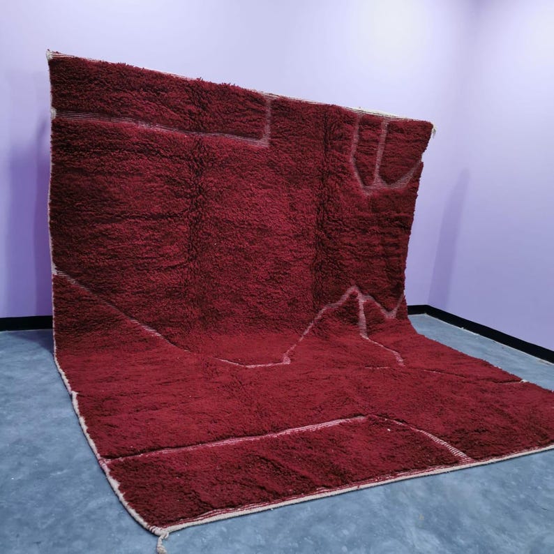 May include: A large, rectangular, burgundy-colored rug with a thick, textured pile. The rug features a geometric design with white lines outlining shapes. The rug is displayed on a gray surface against a purple wall.