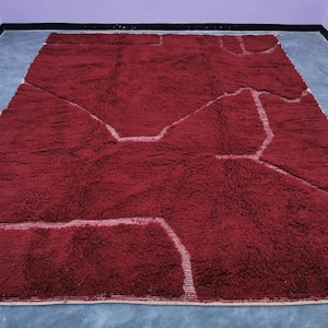 May include: A large, rectangular, burgundy-colored rug with a thick, textured pile. The rug features a geometric design with white, linear accents that create a contrasting pattern across the surface. The rug is suitable for a living room or bedroom.
