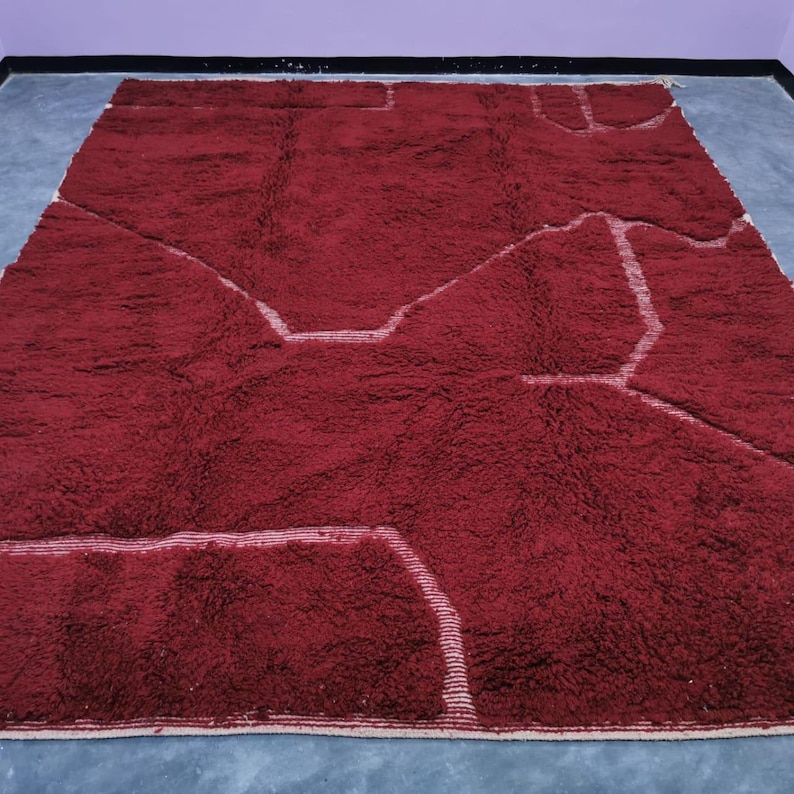 May include: A large, rectangular, burgundy-colored rug with a thick, textured pile. The rug features a geometric design with white, linear accents that create a contrasting pattern across the surface. The rug is suitable for a living room or bedroom.