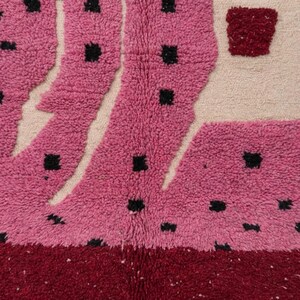 K&ouml;nnte beinhalten: Nahaufnahme eines strukturierten Teppichs mit lebendigem Design. Der Teppich weist eine Kombination aus Rosa, Creme und Burgunderfarben auf. Die rosafarbenen Abschnitte haben schwarze quadratische Akzente und das Design hat ein geometrisches Muster.