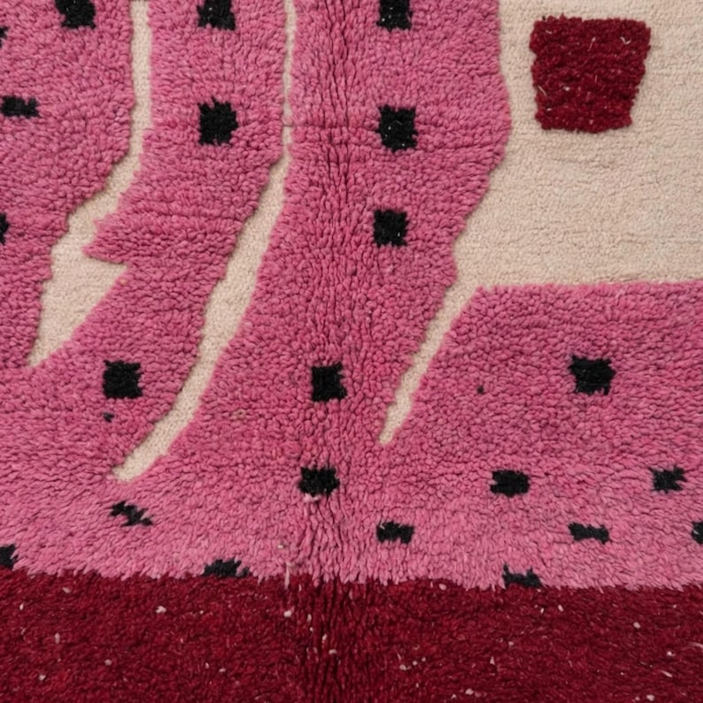 K&ouml;nnte beinhalten: Nahaufnahme eines strukturierten Teppichs mit lebendigem Design. Der Teppich weist eine Kombination aus Rosa, Creme und Burgunderfarben auf. Die rosafarbenen Abschnitte haben schwarze quadratische Akzente und das Design hat ein geometrisches Muster.