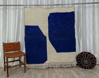 May include: A large, rectangular rug with a geometric design. The rug features a bold color block pattern of cream and deep blue. The rug is hanging on a metal rod against a white curtain backdrop. A brown leather chair and a round ottoman are in the foreground.