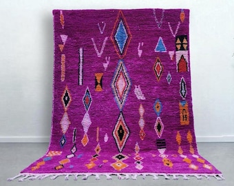 May include: A vibrant magenta area rug featuring a series of geometric diamond patterns in contrasting colors including blue, orange, black, and white. The rug has a textured pile surface and is finished with a row of white fringe along the bottom edge.