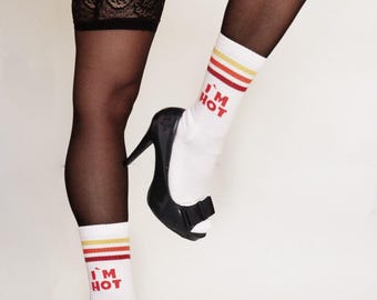 Pode incluir: Um par de meias brancas com listras vermelhas e o texto "I'M HOT" em cada meia. As meias são usadas com saltos altos pretos e meias pretas.