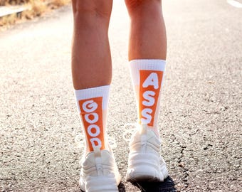 May include: White athletic shoes and socks with the words "GOOD ASS" printed in orange and white. The socks are visible on the lower legs of a person standing on a paved road.