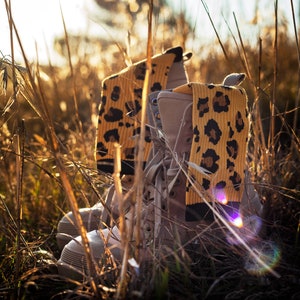 May include: A pair of beige combat boots with yellow and brown leopard print socks. The boots are partially obscured by tall grass.