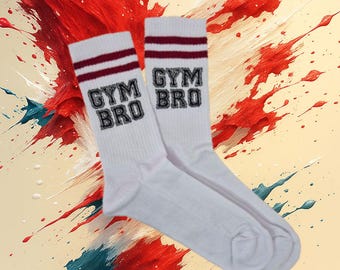 Pode incluir: Um par de meias esportivas brancas com listras bordô e a inscrição "GYM BRO". As meias são exibidas em um fundo de respingos de tinta vermelha, branca e azul. Produto com tema fitness.